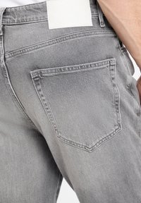 Jeans de denim gris con una vista en primer plano del bolsillo trasero, que presenta una etiqueta de marca de cuero blanca y detalles de costura prominentes.