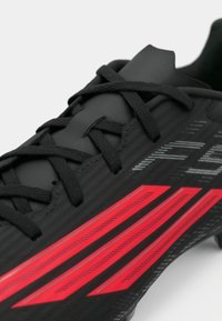 Μαύρο παπούτσι ποδοσφαίρου με ανάγλυφη επάνω επιφάνεια, που διαθέτει τρεις κόκκινες ρίγες και μαύρα κορδόνια. Ο σχεδιασμός περιλαμβάνει διακριτικές γκρι λεπτομέρειες και μοτίβα.