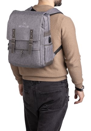 Sac à dos en tissu gris avec un rabat, deux sangles avant et des poches latérales. Comprend une fermeture éclair et une base renforcée pour plus de durabilité.