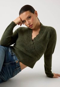 Pulover în culoarea verde măsliniu, cu decolteu sawan și țesătură texturată, având o siluetă ajustată. Asortat cu blugi albaștri ce au un nasture mic.