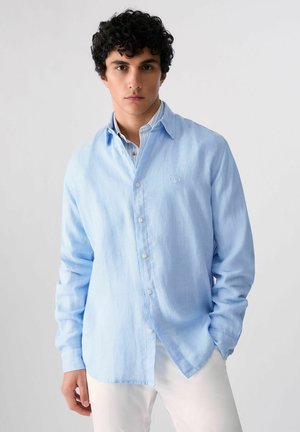 Giovane uomo con capelli scuri e ricci che indossa una camicia azzurro chiaro abbottonata sopra una camicia con colletto a righe e pantaloni bianchi, in piedi davanti a uno sfondo bianco.