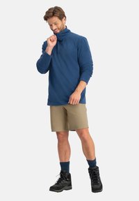 Blauwe fleece trui met een hoge kraag, gecombineerd met beige korte broek, donkere sokken en zwarte wandelschoenen. De stof oogt zacht en heeft een textuur.