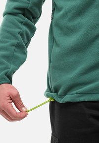 Veste en polaire de couleur vert foncé avec une texture douce. Ourlet ajustable avec un cordon de serrage vert fluo en train d'être tiré. Pantalon noir partiellement visible.