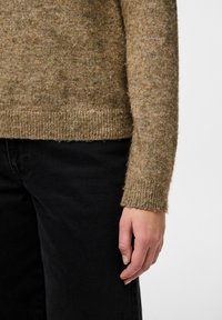 Maglione lavorato a maglia marrone con finitura testurizzata e polsini a costine, abbinato a pantaloni in tessuto neri. La mano si appoggia delicatamente sul tessuto.