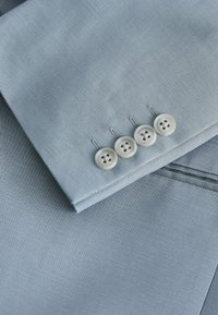 Manche de costume bleu clair avec quatre boutons blancs alignés en rangée près du poignet, montrant la texture du tissu et les détails de couture.
