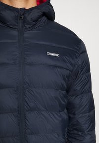 Námořnická puffer bunda s předním zipem, vysokým límcem a kapucí. Interiér je podšitý červenou látkou a na hrudi je umístěno logo.