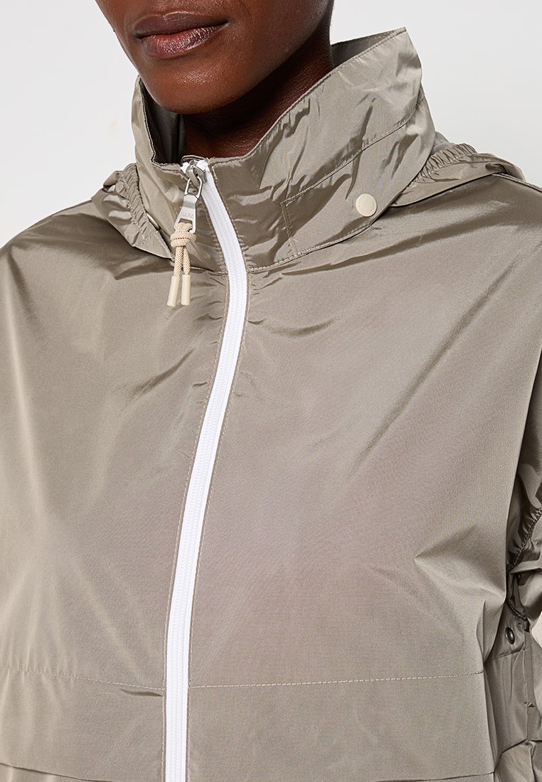 Giacca windbreaker beige chiaro con colletto alto, chiusura frontale con zip e cordini regolabili sul cappuccio con hardware bianco opaco.