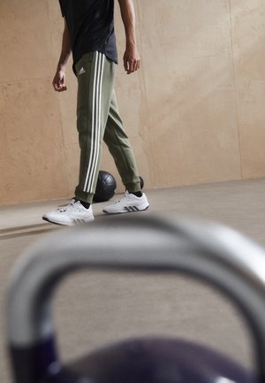 Osoba ubrana w zielone spodnie dresowe Adidas i białe trampki stojąca obok czarnej piłki lekarskiej na siłowni, z kettlebellem na pierwszym planie.