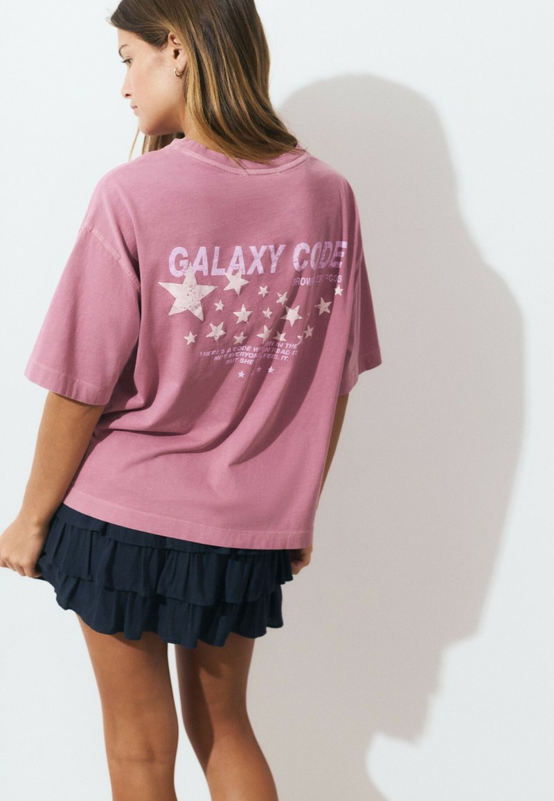 Pink oversized t-shirt with white graphic text and star design on the back; paired with a black ruffled skirt. Smooth cotton texture.