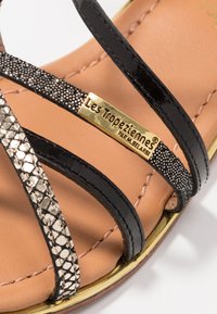 Sandal nera con cinturini e accenti metallici testurizzati, arricchita da una placca con logo dorato. Presenta una soletta liscia color tan e cuciture decorative.