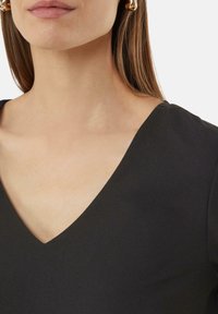 Zwarte V-hals kledingstuk met een gladde, matte textuur. Heeft een aansluitend ontwerp en is gecombineerd met een gouden oorbel in de vorm van een ring.