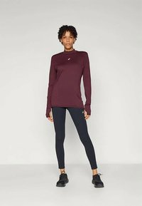 Bordo majica dugih rukava s visokim ovratnikom, koja sadrži mali logo. Uparena s uskim crnim tajicama i crnim sportskim cipelama. Glatka tekstura.