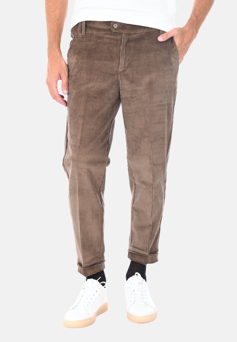 Pantaloni in corduroy marroni con righe verticali, dotati di chiusura con bottone, tasche laterali e risvolti. Indossati con scarpe da ginnastica bianche.