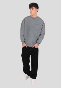 Sivá mikina s rebrovanými manžetami a lemom, s jemným logom v rovnakej farbe; doplnená o čierne voľné nohavice a biele tenisky.