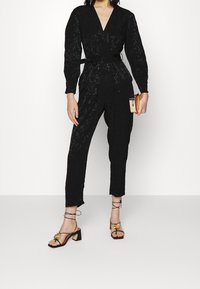 Svart spets jumpsuit med V-ringning, långa ärmar och knytning i midjan. Matchas med svarta klackskor med remmar och ett guldaccent på tån.