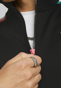 Svart zip-jacka med en rosa dragkedja, med ett texturerat tyg och en vit skjorta under. En silverring syns på handen.