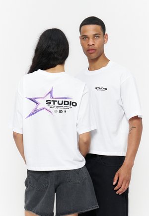 Deux modèles portent des t-shirts courts blancs avec le texte « STUDIO » ; l’un montre le logo avant, l’autre montre le dos avec un graphisme étoile et un slogan.