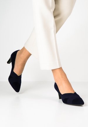 Granatowe zamszowe szpilki z wydłużonym noskiem, z krzyżującym się paskiem i 5 cm blokowym obcasem. Noszone z jasnymi spodniami.