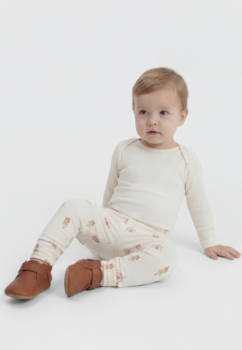 Bambino piccolo con capelli castano chiaro seduto, indossa una maglietta a costine crema a maniche lunghe, pantaloni crema a fantasia e morbide scarpe marroni per bambini su sfondo bianco.