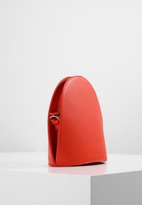 Czerwona sk�órzana torebka z zaokrąglonym wierzchem i widocznym zamkiem błyskawicznym z boku, stojąca w pionie na białym cokole na jednolitym szarym tle.