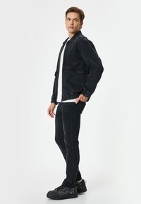 Chaqueta de mezclilla negra con puños elásticos y dos bolsillos frontales, combinada con jeans negros y zapatillas negras texturizadas. Camiseta blanca lisa debajo.