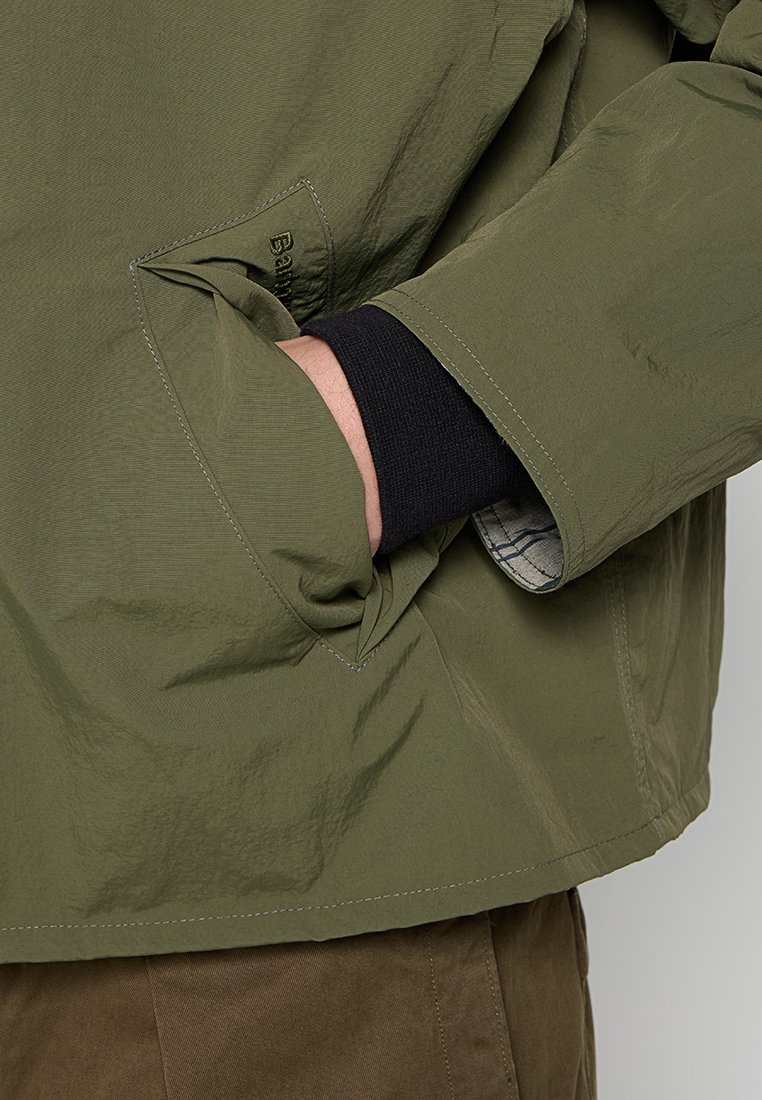 Olijfgroene jas met een gestructureerd oppervlak, voorzien van een zak en een zichtbaar zwart manchet. Eenvoudig ontwerp met minimalistische stikdetails.
