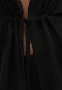 Μαύρο ένδυμα με συγκεντρωμένο ύφασμα και δεμένους ιμάντες που αποκαλύπτουν διαφανές υλικό και δαντέλα από κάτω.