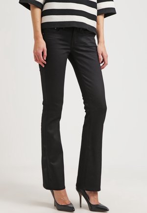 Femme portant un pantalon noir slim bootcut, des talons aiguilles noirs et un haut à manches courtes rayé noir et blanc, debout devant un fond uni.