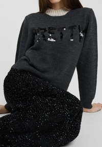 Donna che indossa un maglione grigio scuro con la parola "PRETTY" in paillettes e pantaloni neri lavorati, seduta con una mano appoggiata a terra.