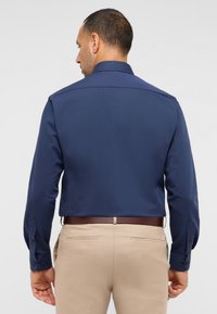 Maglietta a maniche lunghe blu navy con colletto classico, dotata di polsini con bottone e texture liscia, abbinata a pantaloni beige chiaro.
