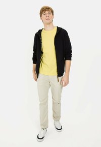 Sort zip-up hoodie med gul t-shirt, beige bukser og hvide sneakers. Hoodie har logodetaljer; glatte stofteksturer i hele. 