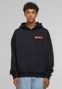 Crna hoodie s kengurskom džepom, natpis "DANI PRIJE" u crvenom na lijevoj strani prsnog dijela, rebrasti rukavi i donji rub, izrađena od mekanog pamučnog materijala.