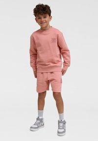Rosa sweatshirt och shorts-set i mjukt material, med en liten logotyp på bröstet, parat med grå höga sneakers och strumpor.