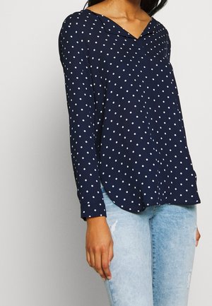 Marineblauwe blouse met lange mouwen en witte polka dots, een V-halskraag en een afgeronde zoom. Gecombineerd met lichtgewassen, beschadigde jeans.