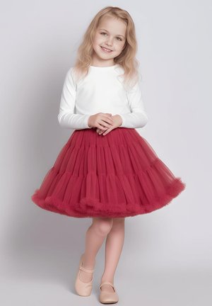 Młoda dziewczynka uśmiechająca się, ubrana w białą bluzkę z długim rękawem, czerwoną spódnicę z warstw tiulu oraz beżowe buty typu Mary Jane, stojąca na jednolitym tle.