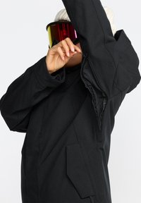 Schwarze Skijacke mit einem Reißverschlussbelüftungsöffnung unter dem Arm, mit einem strukturierten Innenfutter und einer Vorderseite-Tasche.