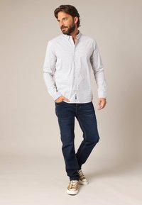 Heren overstijd overhemd in het wit met een klein blauw ruitpatroon, gecombineerd met donkerblauwe jeans en bruine sneakers met witte zolen.