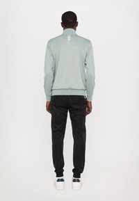 Jachetă sport de culoare verde deschis cu guler în picioare și accente reflexive, asortată cu pantaloni jogger negri și adidași albi cu detalii verzi.