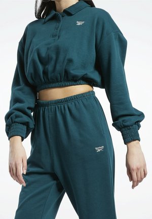 Kobieta ubrana w turkusowy, krótki, długi rękaw top Reebok z elastycznym pasem i dopasowane, wysokie spodnie dresowe z elastycznymi mankietami.