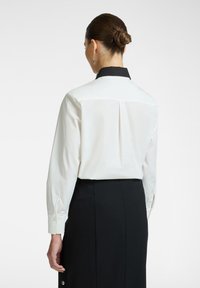 Camicia bianca a maniche lunghe con colletto nero, caratterizzata da una piega sul retro e polsini con bottoni, abbinata a una gonna nera. Design semplice e classico.