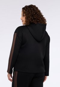 Sudadera con capucha negra con un bolsillo frontal, presenta rayas verticales marrones en las mangas, hecha de un material liso y suave, con capucha ajustable con cordón.