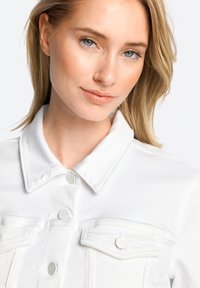 Weißes Denim-Jacket mit durchgehendem Knopfverschluss, großen Brusttaschen und flachem Kragen. Ausgestattet mit dezenten Nähten und runden metallischen Knöpfen.
