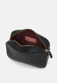 Černá kožená kosmetická taštička s texturovaným povrchem, zlatým zipem a bordovým vnitřkem. Uvnitř má štítek s označením značky.