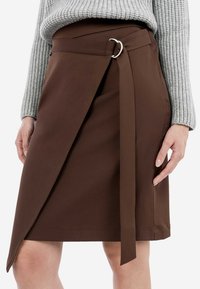 Hnědá zavinovací sukně vyrobená z hladkého materiálu, s bočním uzlem a stříbrným kruhovým akcentem. Stylizováno s šedým žebrovaným svetrem.