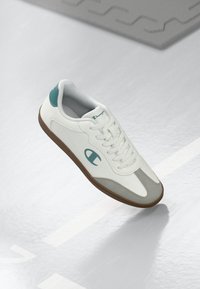 Hvit sneaker med lysegrå detaljer, med en teksturert lærøvre, rund t�å, teal-bekledning på hælen og gummisåle.