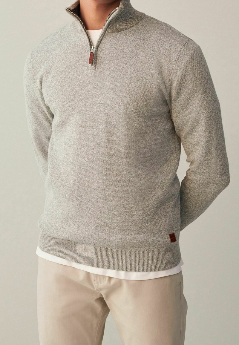 Hellgrauer Pullover mit Reißverschluss, geripptem Kragen und Bündchen, mit einem kleinen braunen Lederetikett am Saum. Tragen über einem weißen Hemd.