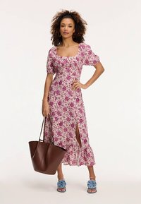 Vestido midi floral con mangas abullonadas, escote cuadrado y apertura lateral, con estampados en tonos rosa y púrpura. Bolso tote marrón y sandalias azules con volantes.