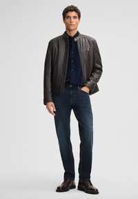 Braune Lederjacke mit gerippten Schultereinsätzen, navyblaues Hemd, dunkelblaue Jeans und braune Lederschuhe. Einfaches, elegantes Design.