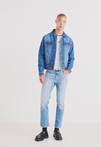 Lys blå denimjakke med to frontlommer og knappelukking, brukt over en hvit T-skjorte, sammen med lyseblå jeans og svarte sko.