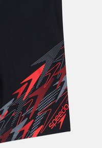 Μαύρο ύφασμα με κόκκινο και γκρι αφηρημένο μοτίβο βέλους και τη λέξη «Speedo» τυπωμένη με κόκκινα γράμματα στο κάτω δεξί μέρος.